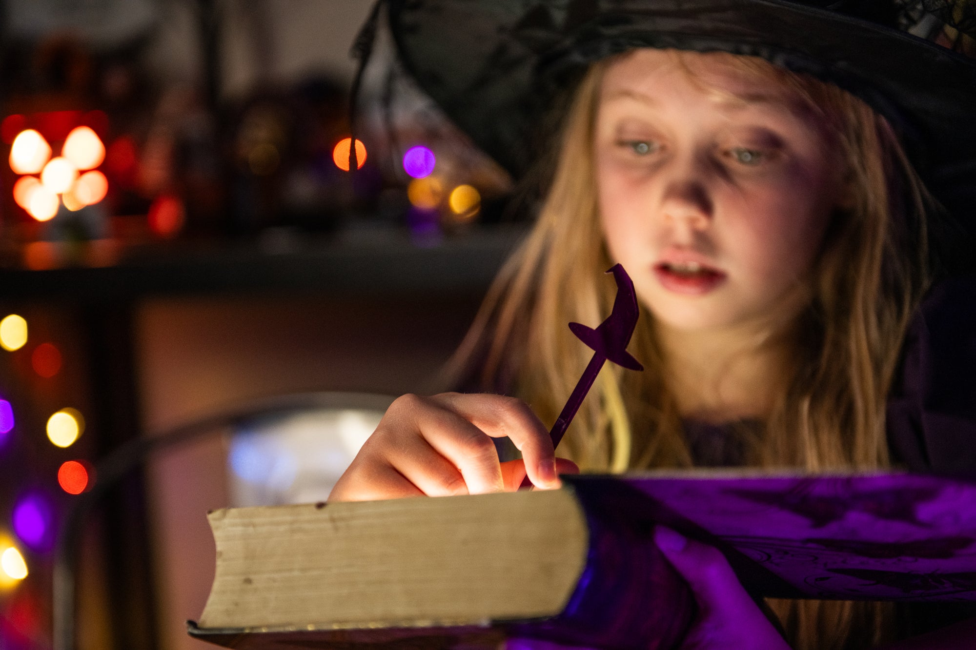 Child in witch costume holding a candle and open book with blurred lights in the background